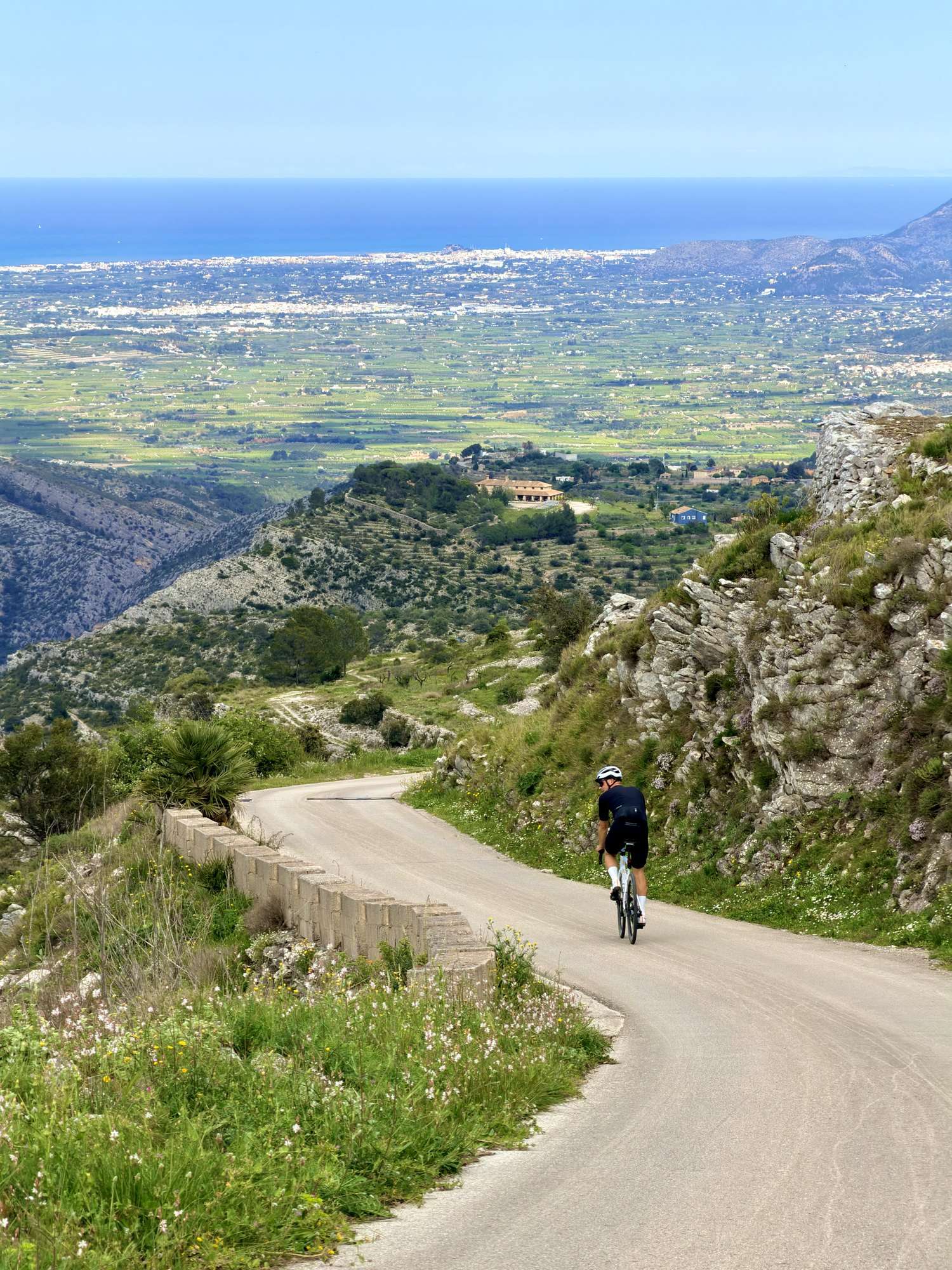 Un cycliste sur route descend une route de montagne sinueuse, avec la vallée et la mer en contrebas, sur la Costa Blanca