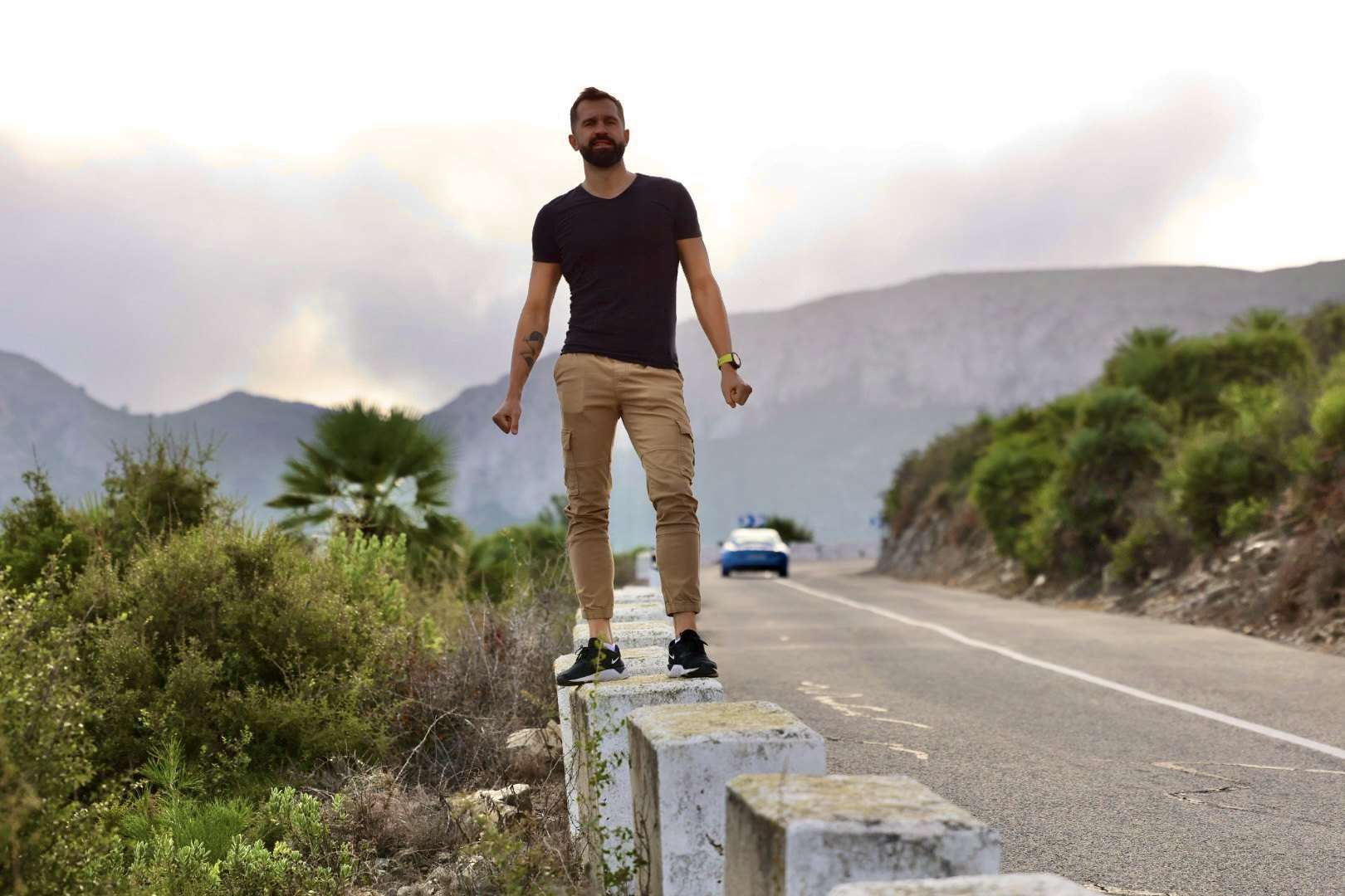 Kamil debout sur une glissière de sécurité au bord d’une route de montagne, avec des pentes et le ciel en arrière-plan