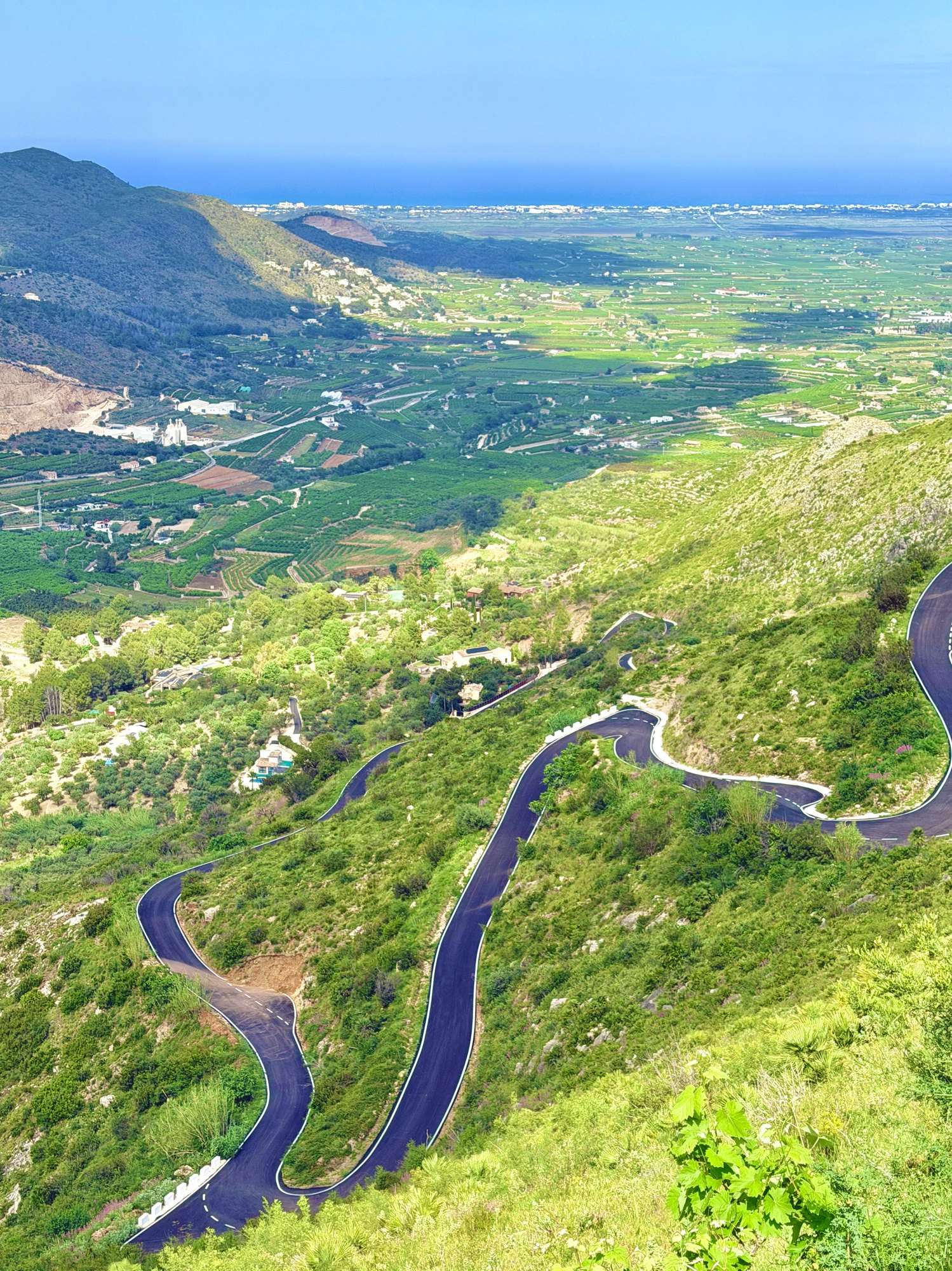 Des virages en épingle sur une colline verte au-dessus d’une vallée, avec la côte de la Costa Blanca en arrière-plan